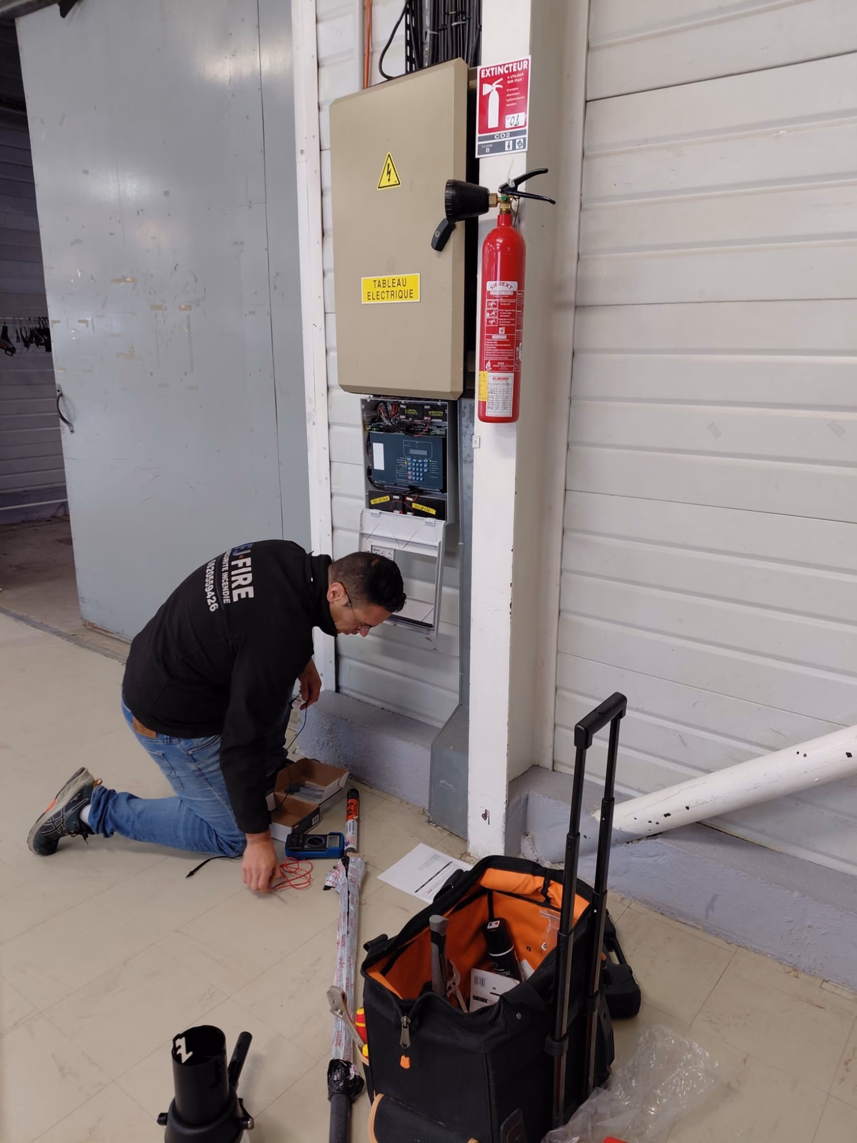 Technicien FJ Fire agenouillé devant la centrale d'alarme incendie ouverte, raccordement électrique en cours, salle de réception à Saint-Seurin-sur-l'Isle